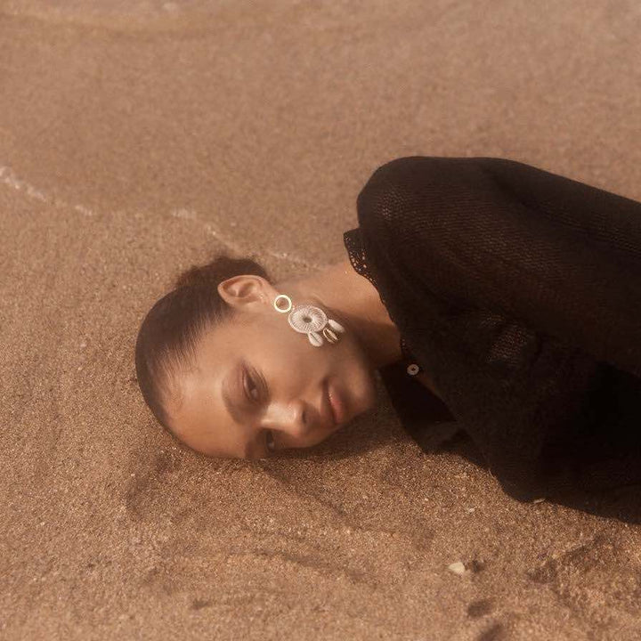 Model lying on beach in black top and cowry shell and gold earrings