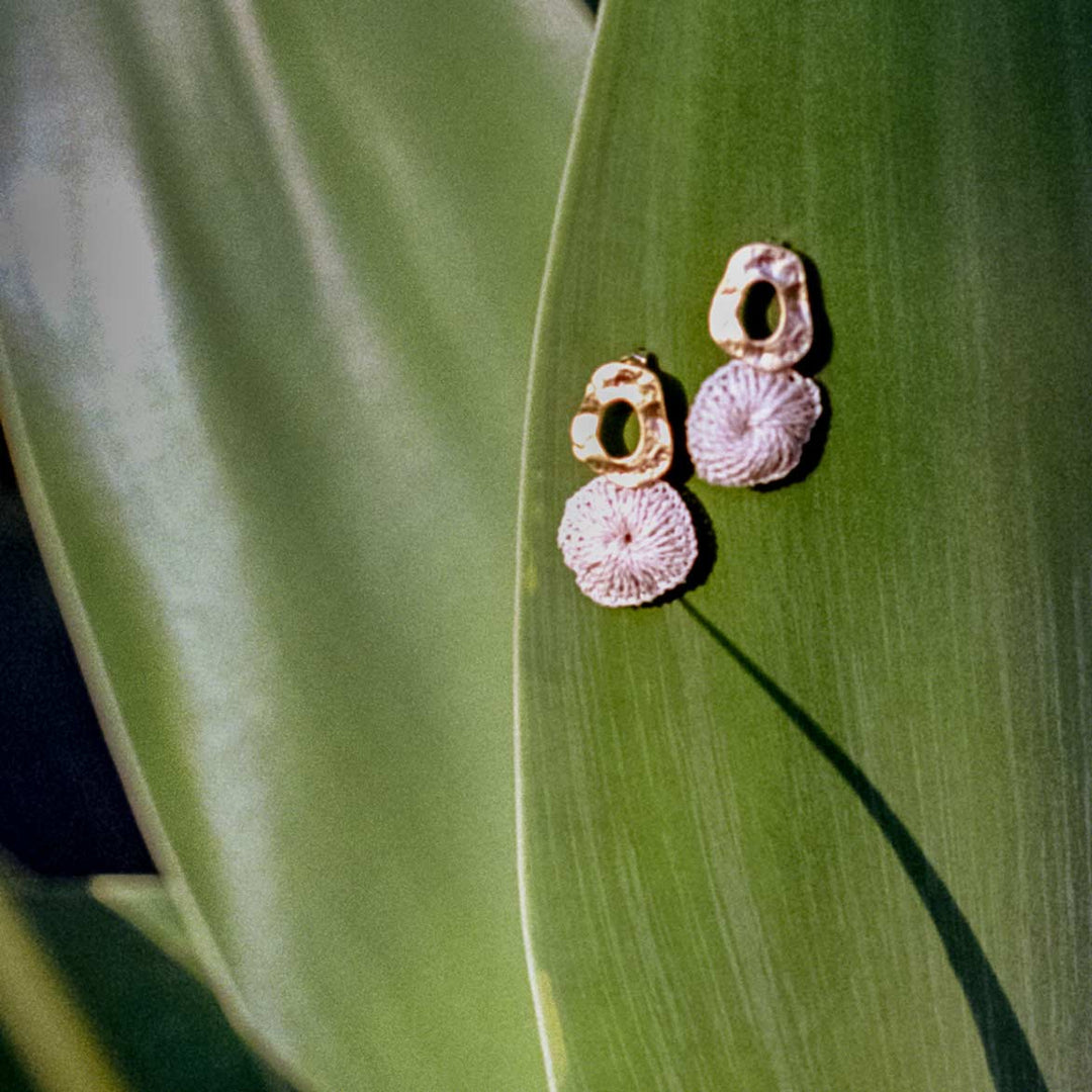 Bilum and Bilas Warp earring with textured gold circle earring and dangling natural fibre disc still life on a green leaf