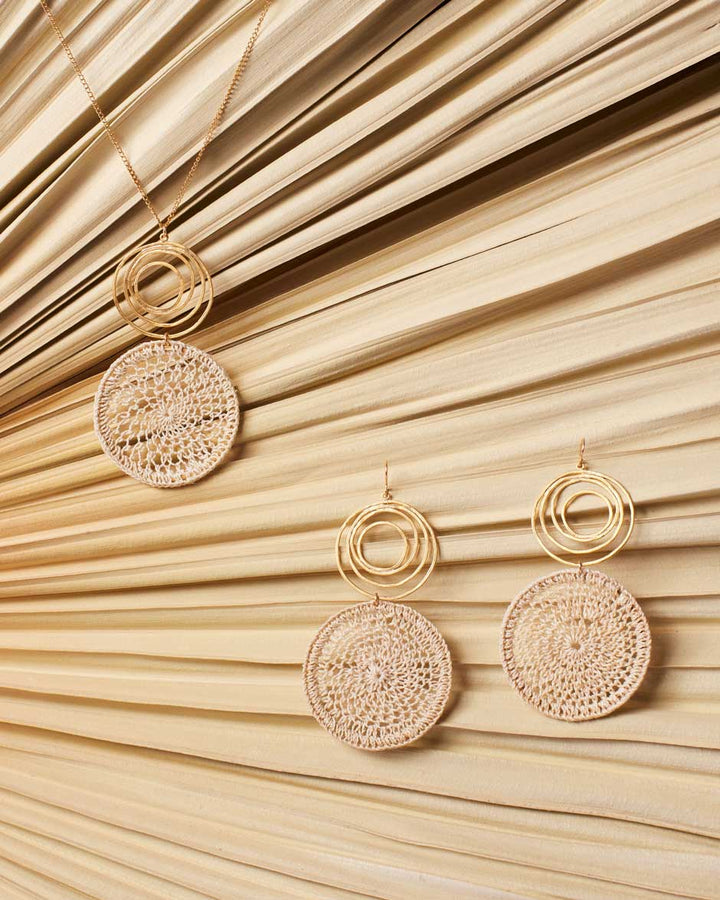 Gold swirl earrings with natural fibre woven disc with matching necklace on a dried palm leaf.