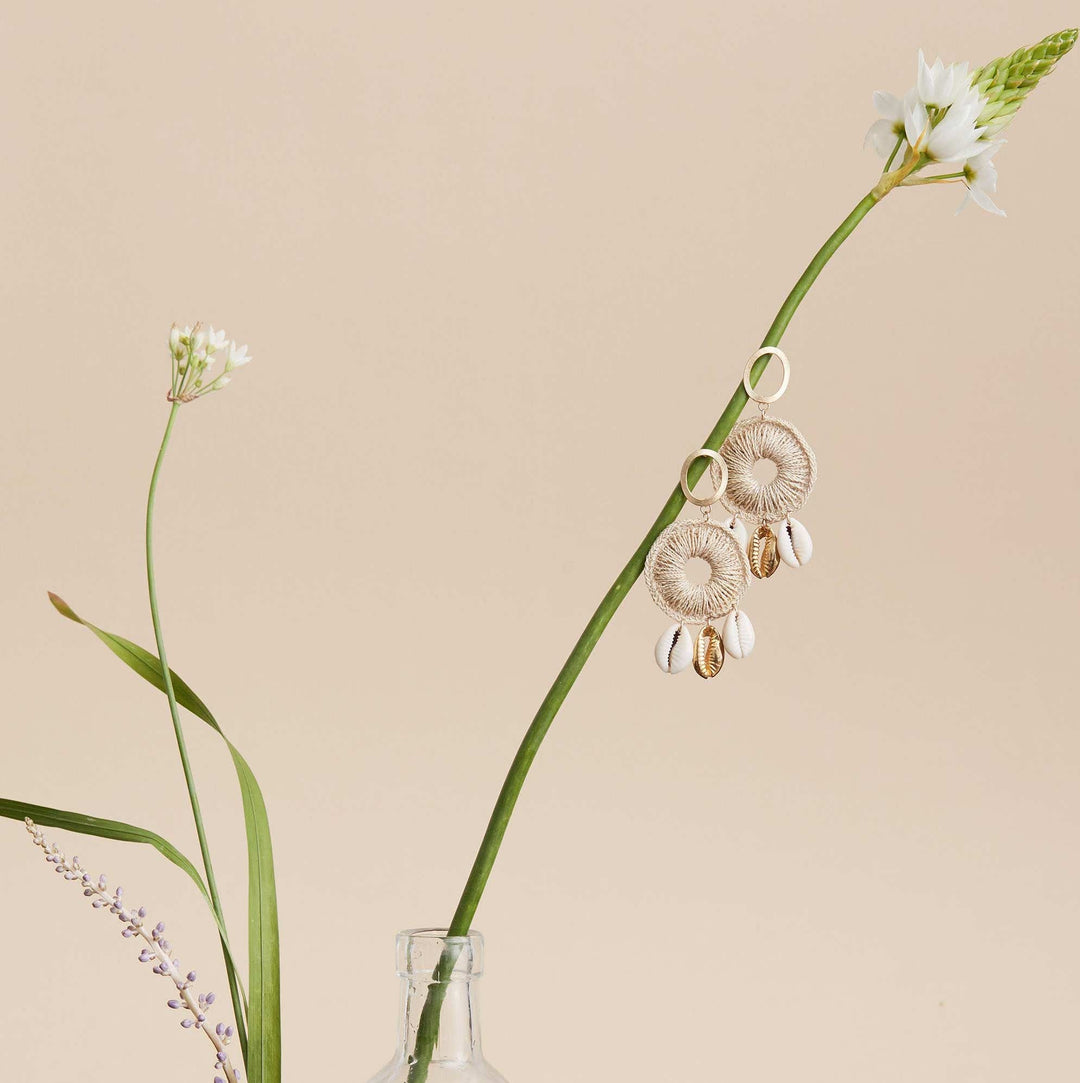 Still life picture of a Dangle statement gold earring with handwoven hoop featuring natural and gold cowry shells