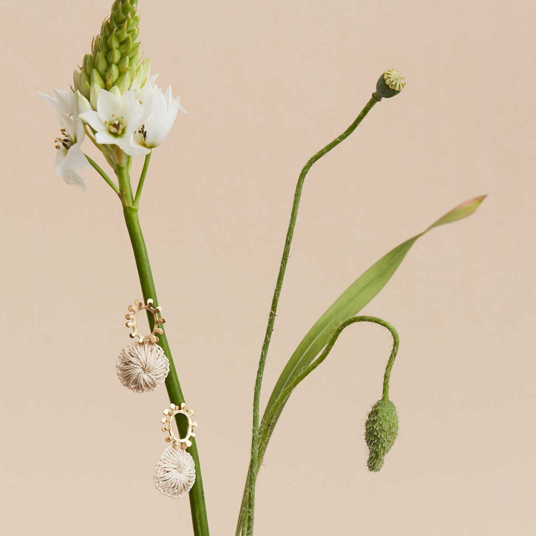 Still life of gold textured sun earrings with natural fibre woven discs displayed on flower stems