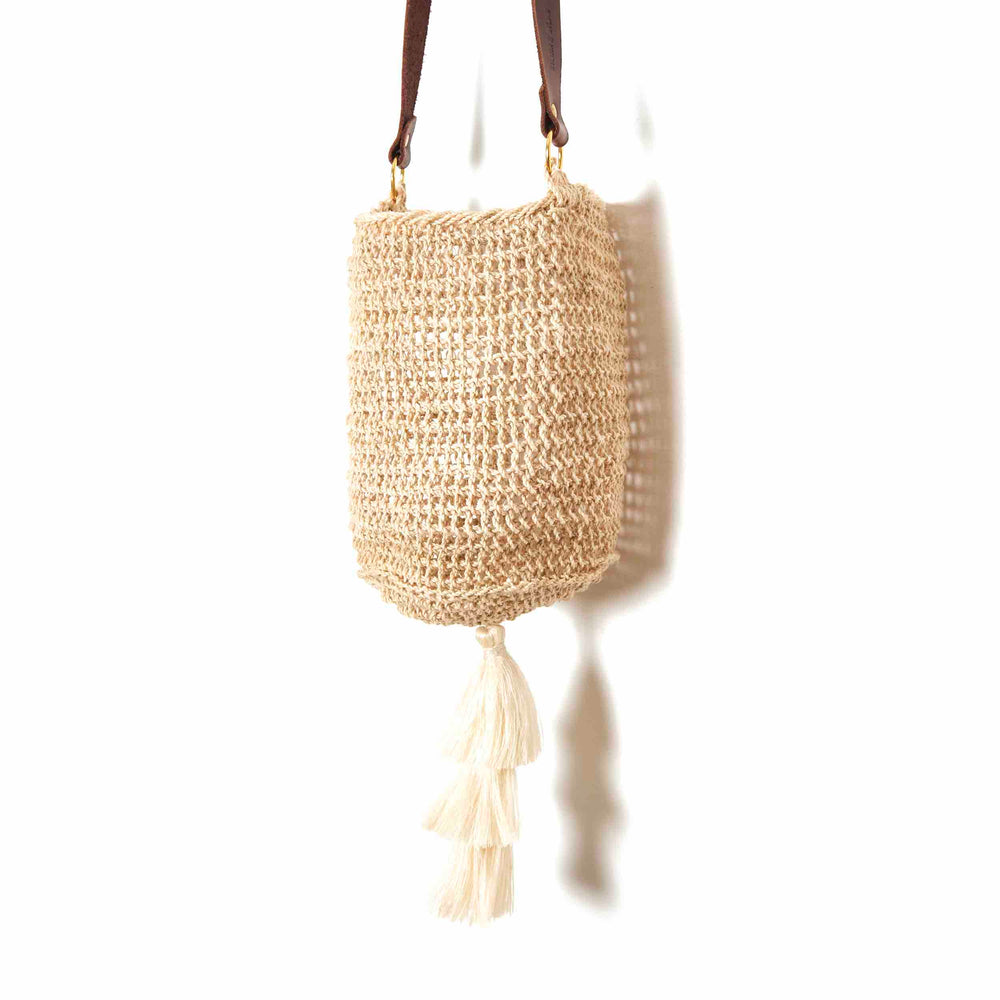 Side angle of natural bucket bilum bag with leather strap and tassel on base hanging on a white background.