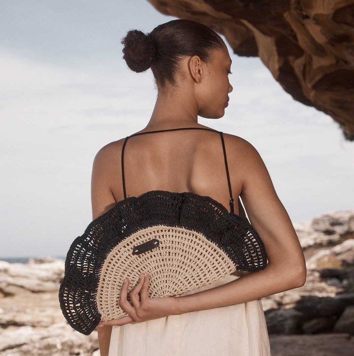 Model holding the Ripple Clutch woven bag in black behind her back standing on rocks.