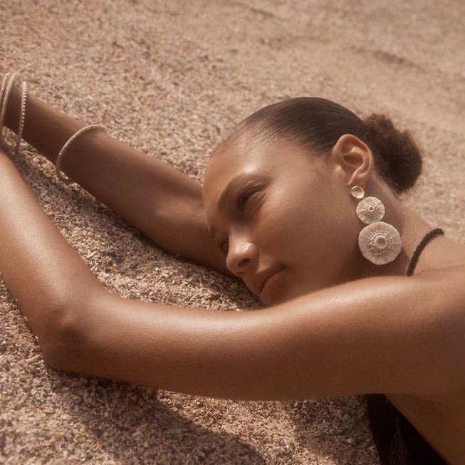 Natural fibre pom pom statement earrings on a model lying in the sand. bilum and bilas