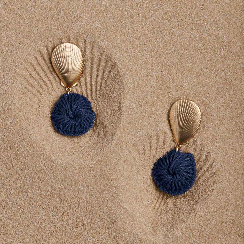 Pair of earrings with blue spherical pendants and gold shell-like tops on a sandy background