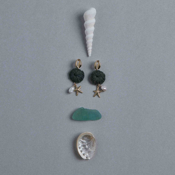 Pair of earrings with starfish and pearl details on a gray background with shells and a piece of green stone.