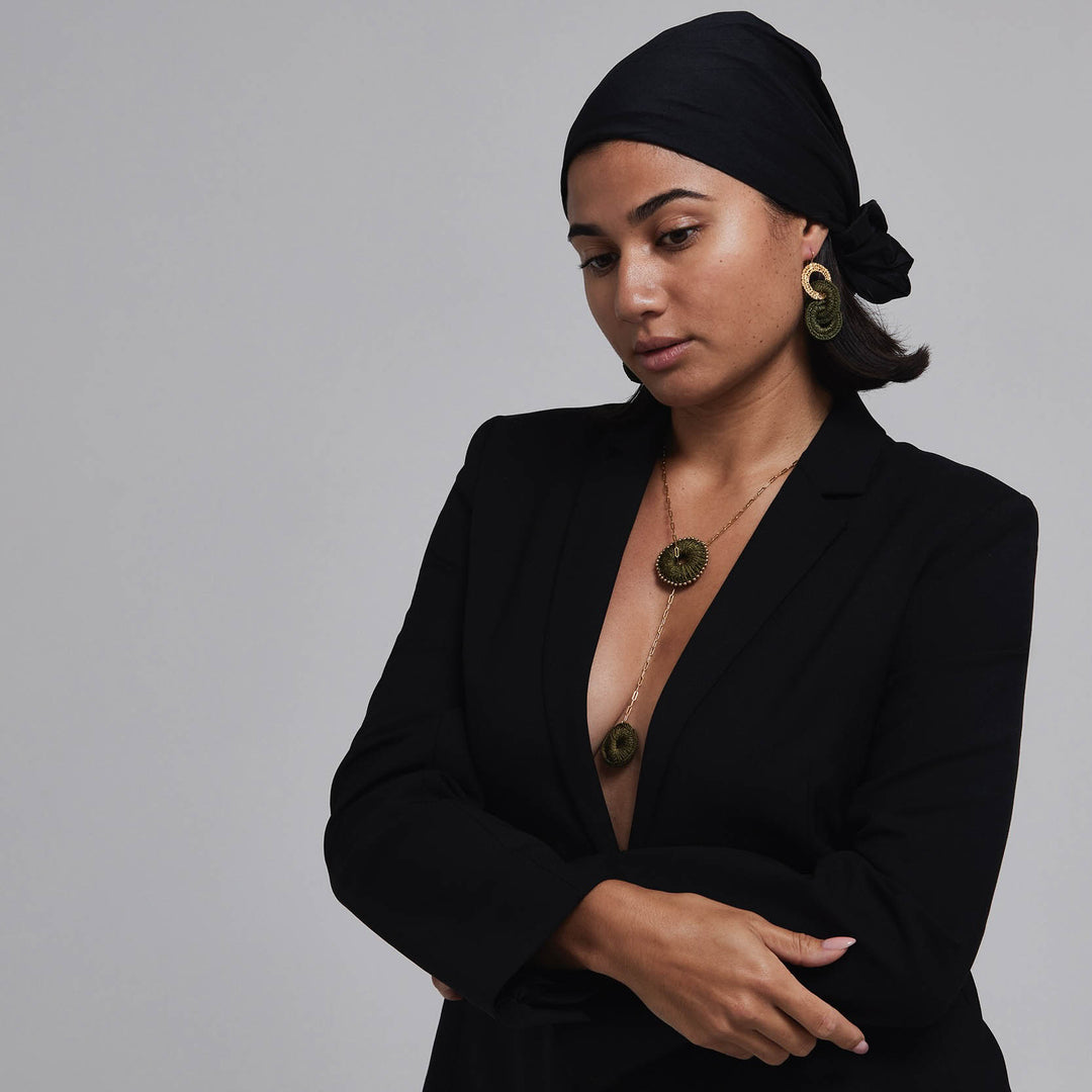 Woman wearing a black blazer and headscarf with gold jewelry on a gray background
