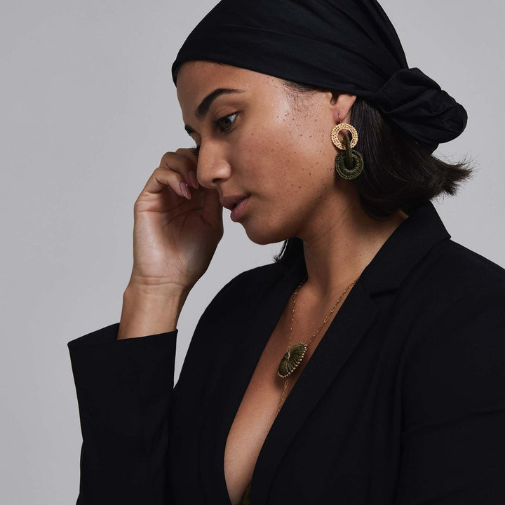Woman wearing a black headscarf and black blazer with gold earrings and necklace against a gray background