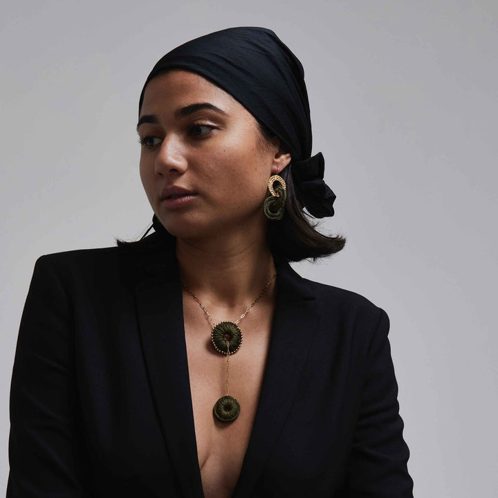 Woman wearing a black headscarf, black blazer, and gold jewelry against a gray background