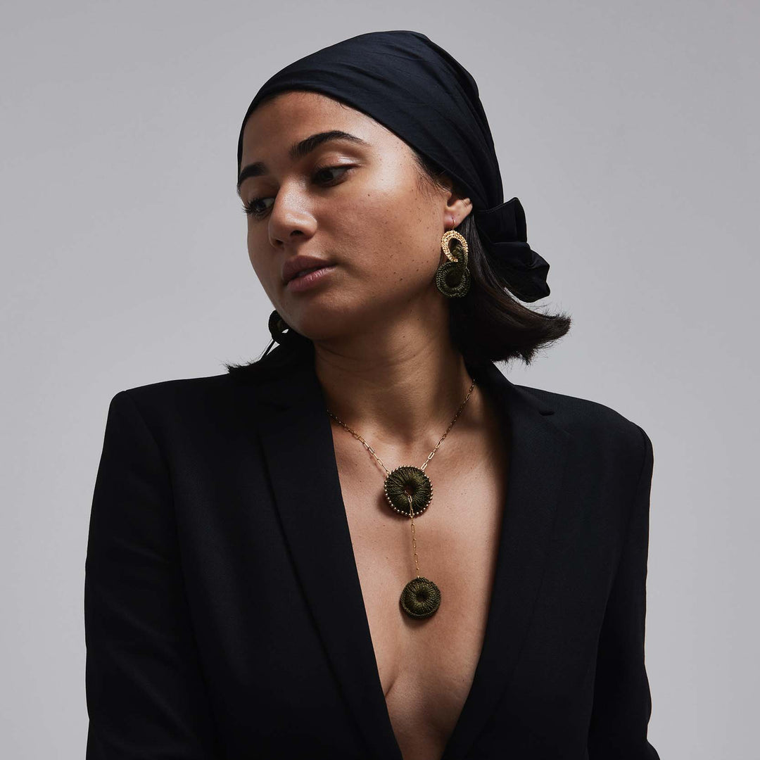 Woman wearing a black blazer and headscarf with a necklace and earrings on a gray background