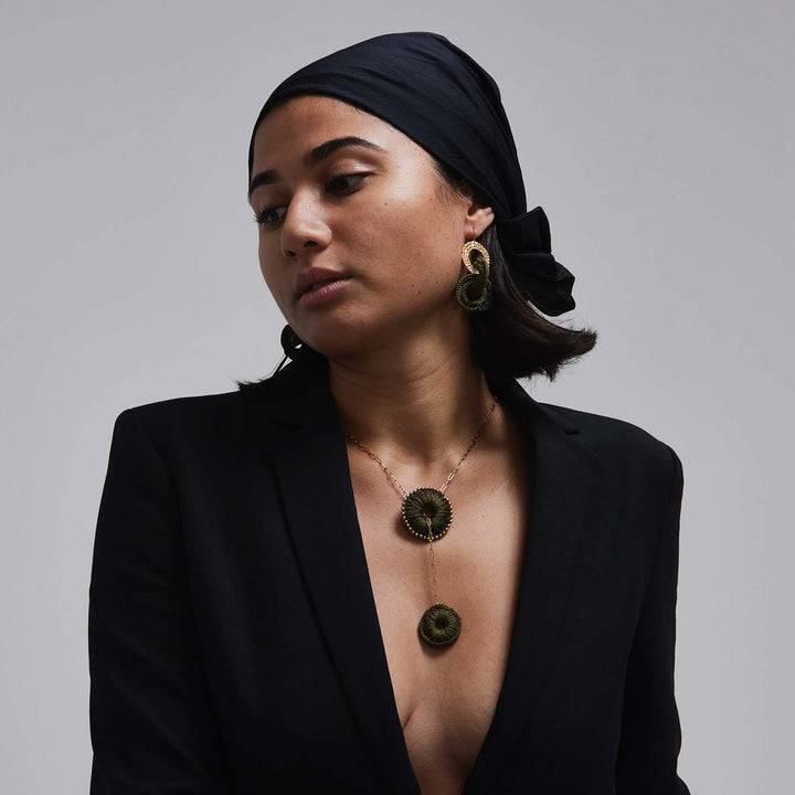 Woman wearing a black blazer and headscarf with a necklace and earrings on a gray background