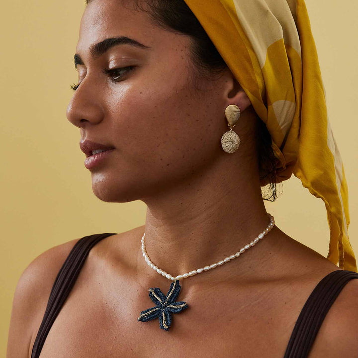 Woman wearing a yellow headscarf, pearl necklace, and earrings against a beige background