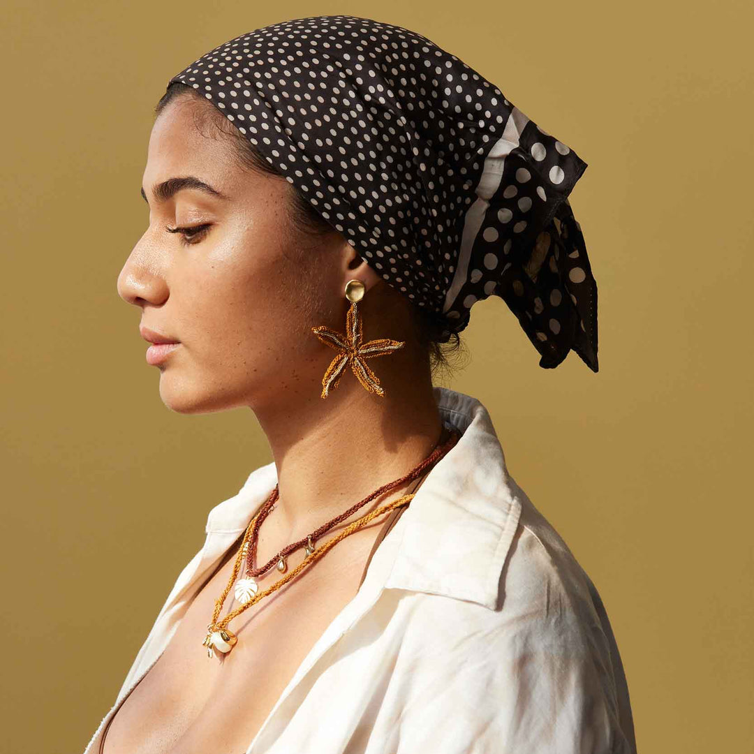 Woman wearing a black polka dot headscarf and gold jewelry against a beige background