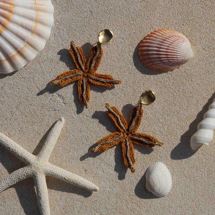Starfish-shaped earrings on a sandy surface with seashells