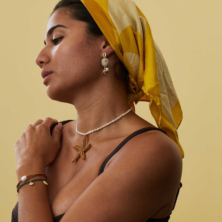 Woman wearing a yellow headscarf, necklace, and earrings against a beige background