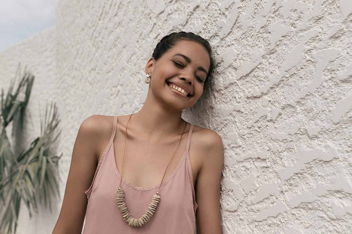 Model laughing land leaning on wall wearing warped gold and natural fibre earrings and necklace Bilum and Bilas