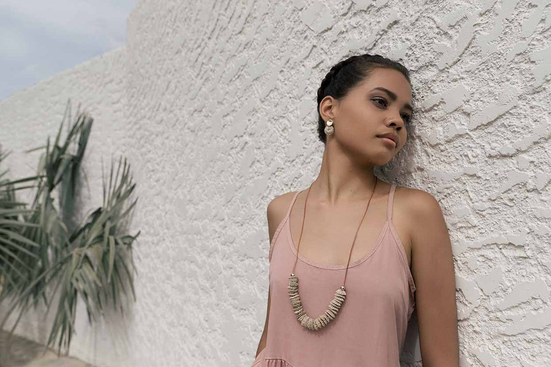 Model leaning on wall wearing Bilum and Bilas warped gold and natural fibre earrings and necklace
