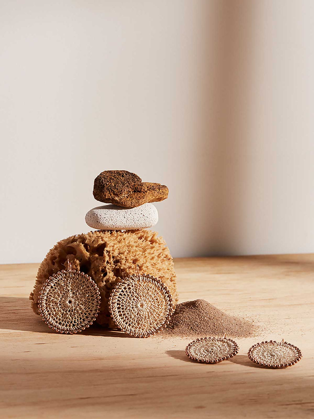Still life with rocks and sand of Bilum and Bilas Sunset earrings. Gold beaded natural fibre handwoven disc earrings.